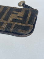 Close-up of a brown and black monogram zip pouch, showing the Fendi Zucca pattern and a gold-tone zipper pull on a light gray background.