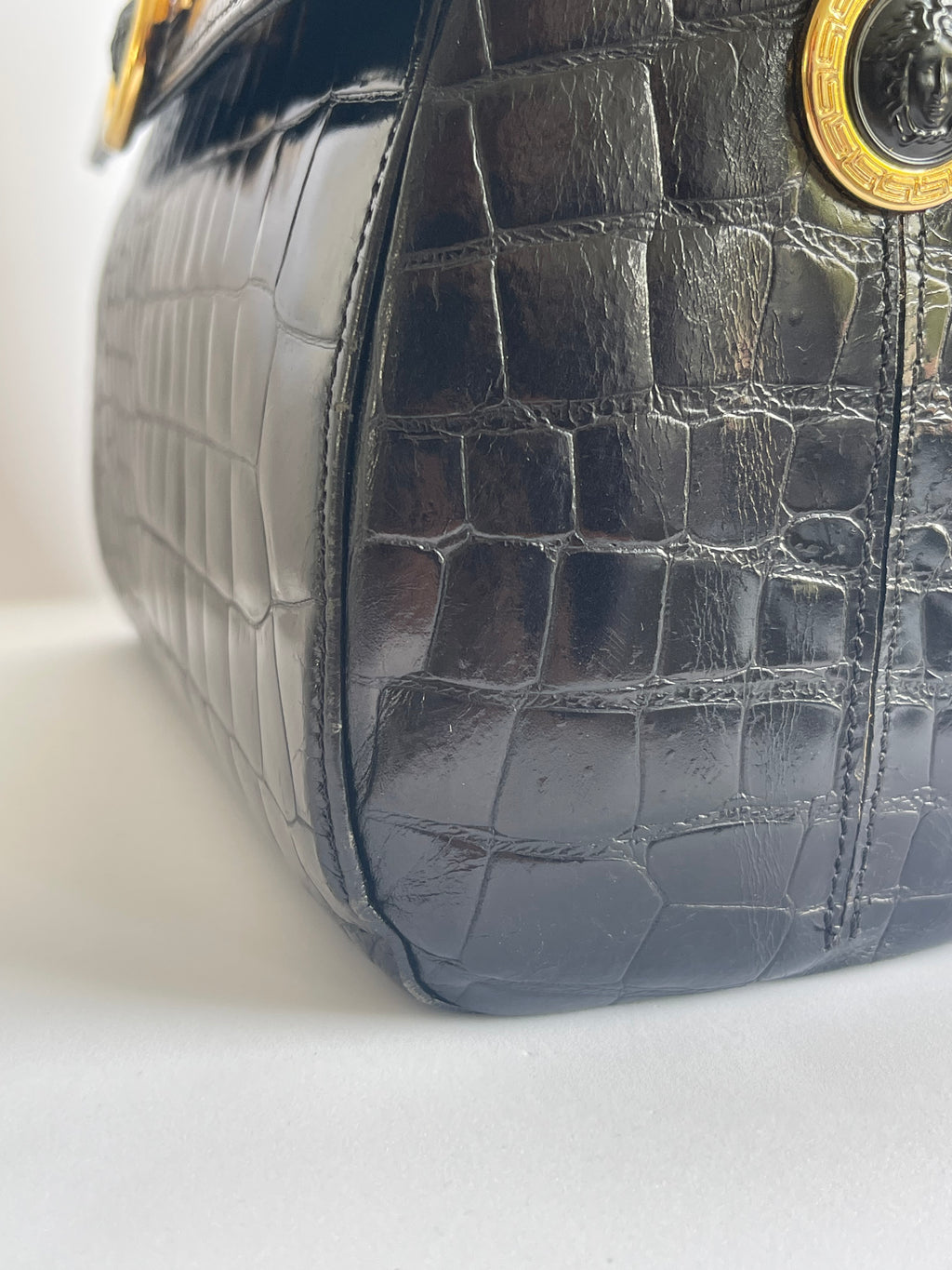 Close-up of a black crocodile-embossed leather handbag corner with gold-tone hardware on a white background.