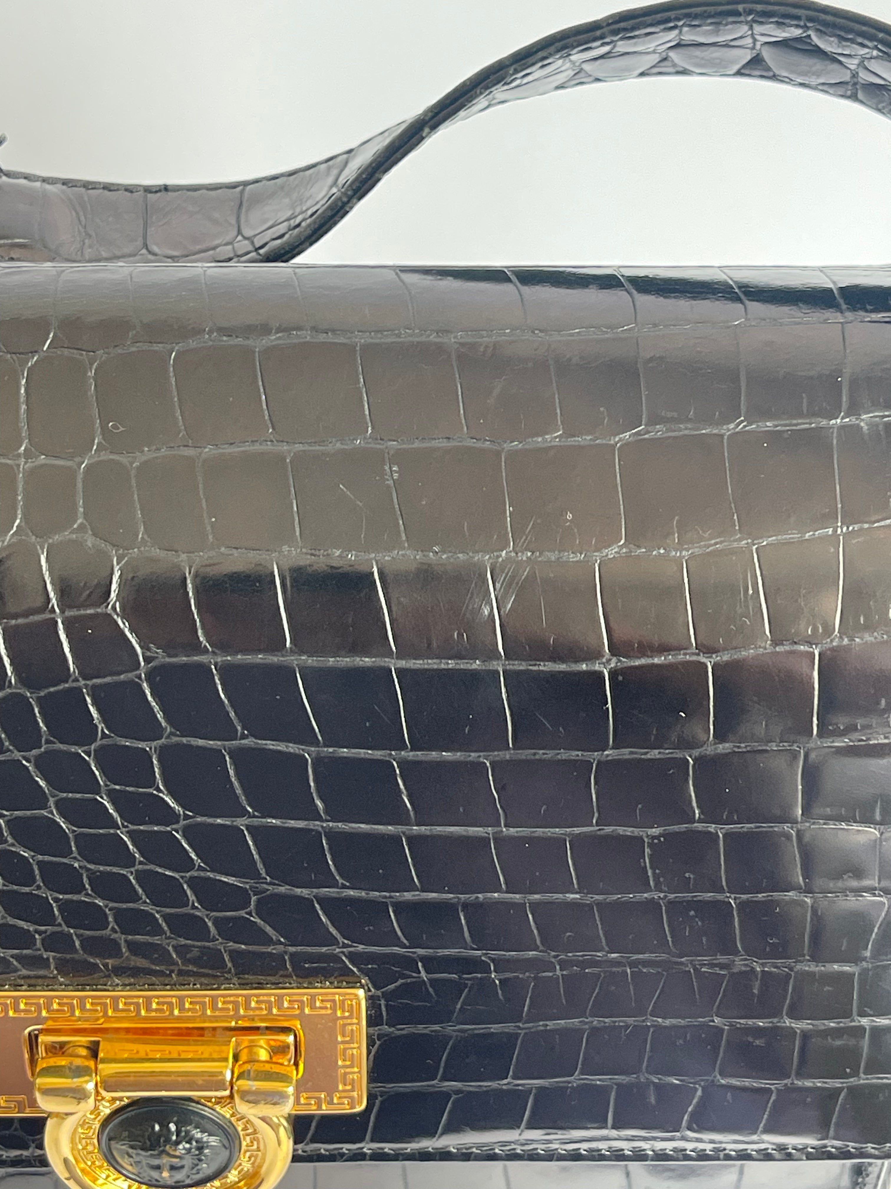 Close-up of a black crocodile-embossed leather handbag with a gold-tone clasp and a light gray background.