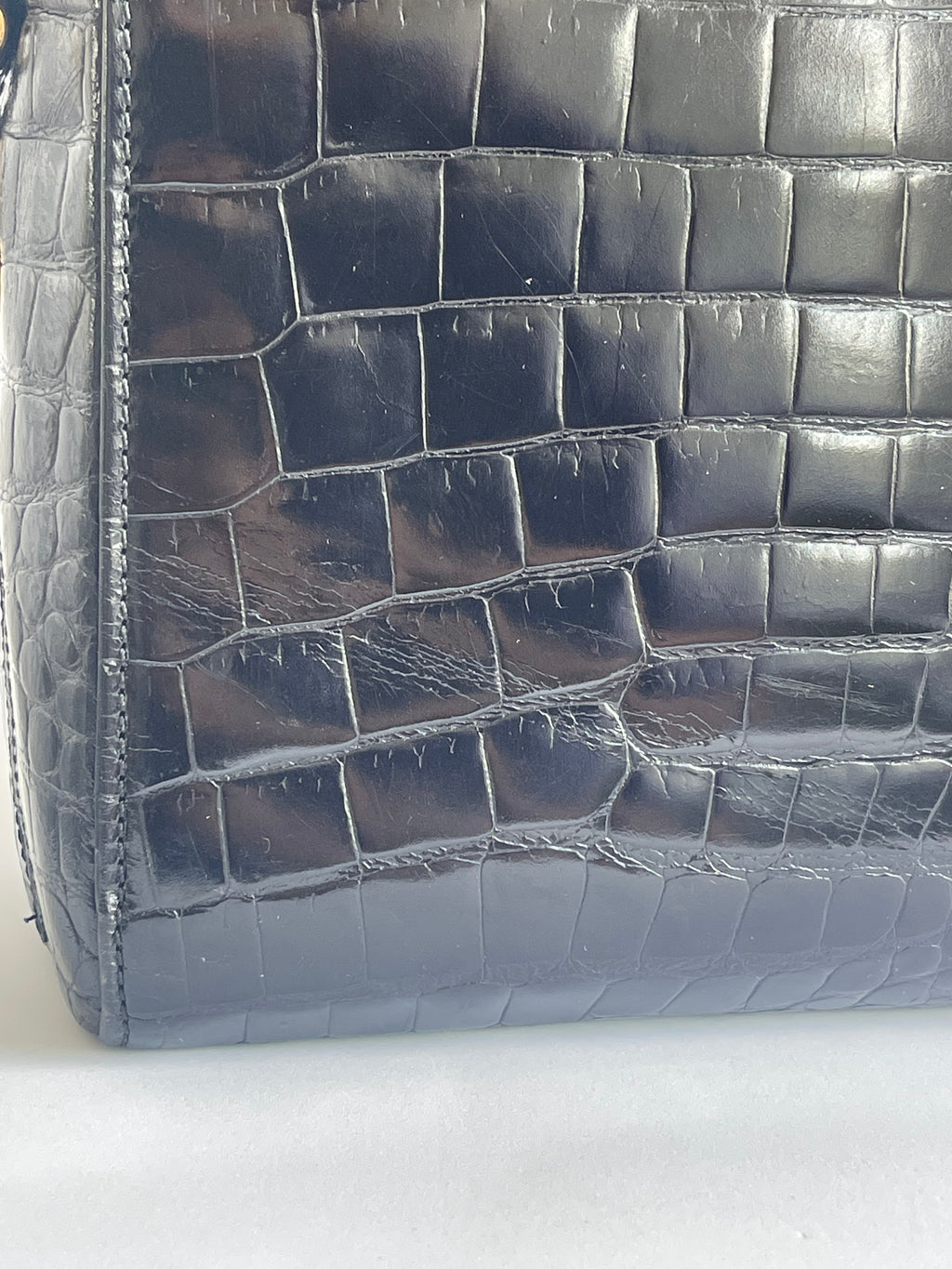 Close-up of a glossy crocodile-embossed leather bag corner in a dark blue-gray color, showing the textured scale pattern and stitching.