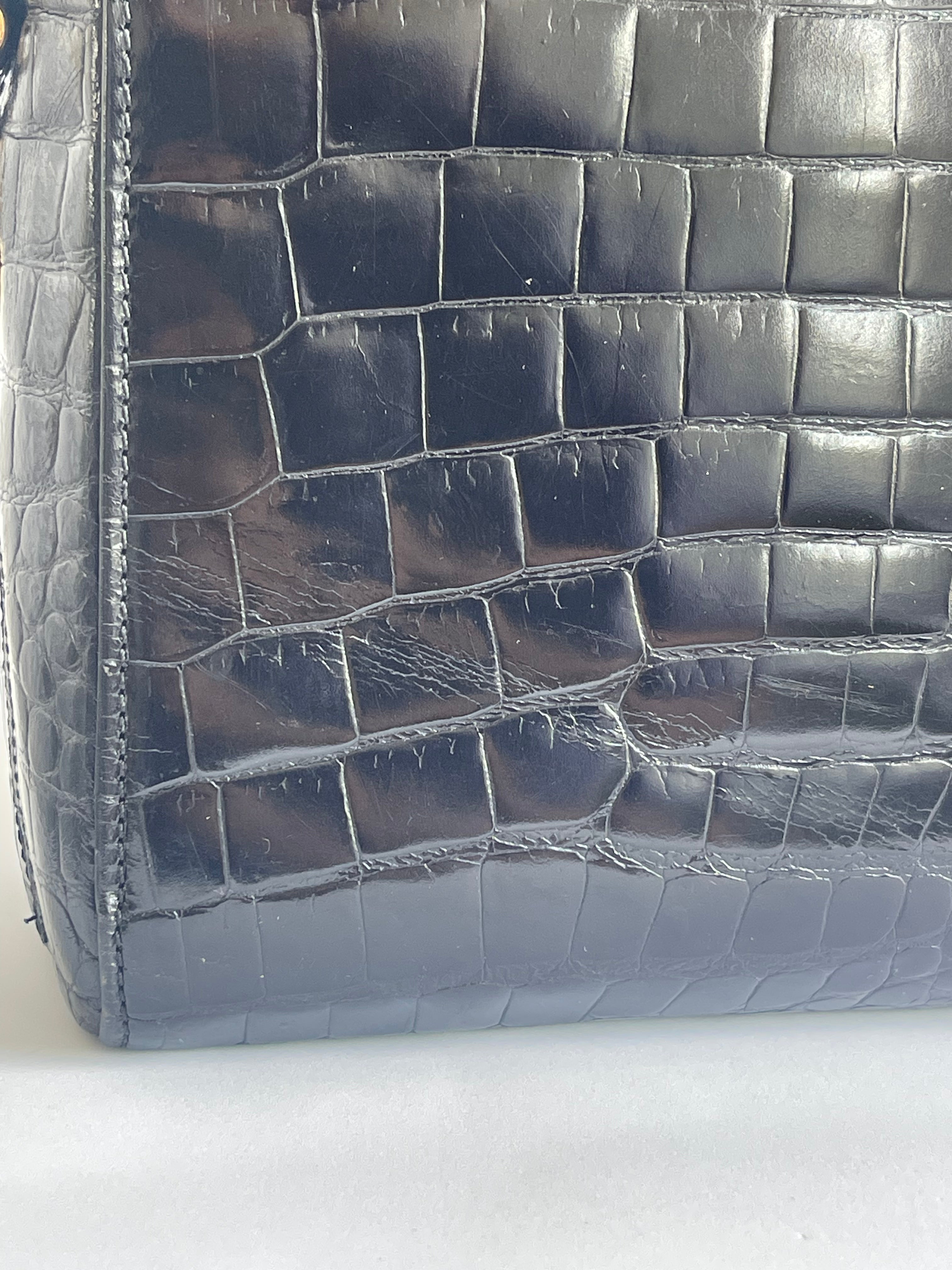 Close-up of a glossy crocodile-embossed leather bag corner in a dark blue-gray color, showing the textured scale pattern and stitching.