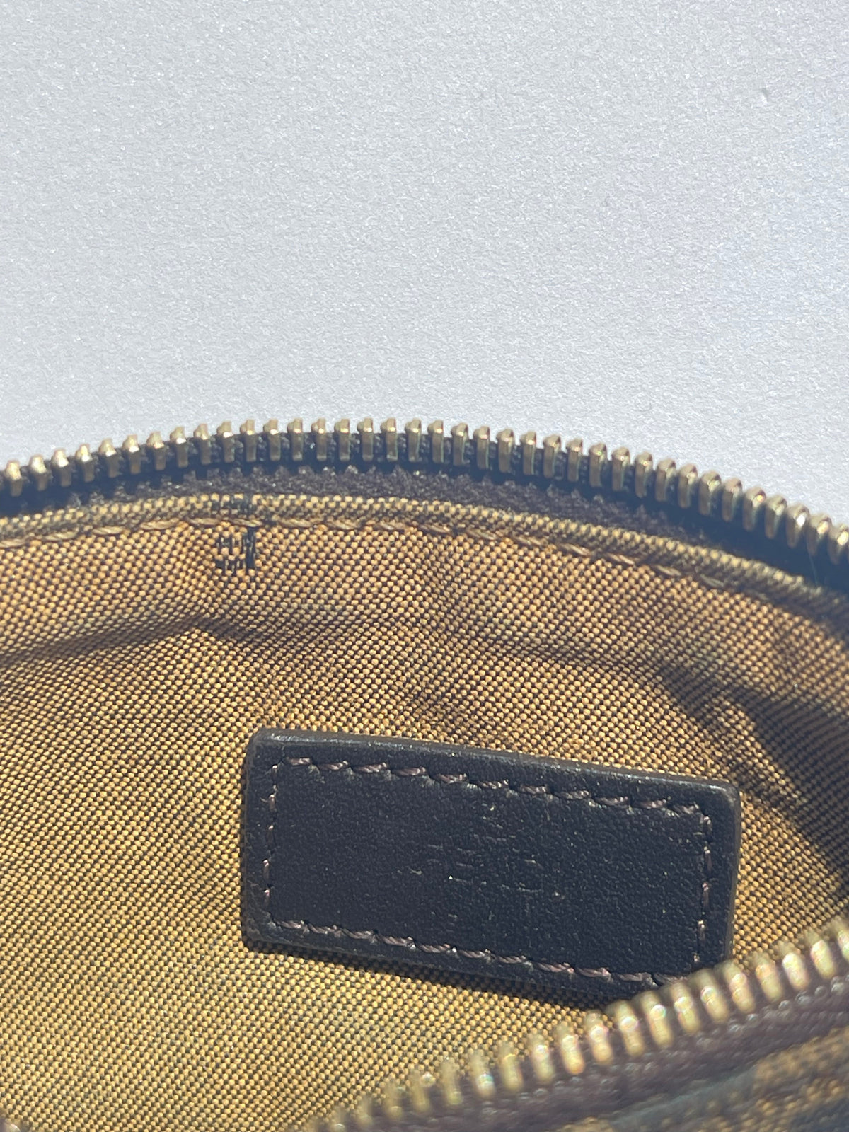 Close-up of the interior of a zippered pouch or bag, showing a tan canvas lining, a black leather brand patch, and a gold-toned zipper.