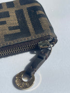 A close-up of a Fendi zip-around wallet in the classic brown and black Fendi Zucca monogram canvas, featuring a dark brown leather pull tab with a gold-tone Fendi logo medallion.