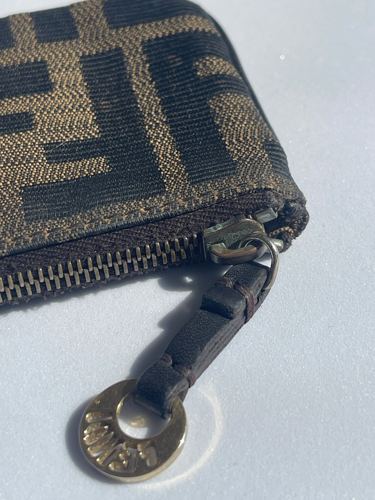 A close-up of a Fendi zip-around wallet in the classic brown and black Fendi Zucca monogram canvas, featuring a dark brown leather pull tab with a gold-tone Fendi logo medallion.