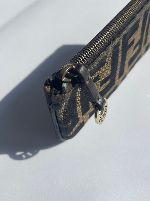 A close-up of a Fendi monogram zipper pouch in black and brown canvas, featuring a black leather pull tab with a silver-tone metal Fendi logo charm.