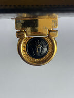 Close-up of a gold-tone metal clasp with a circular black medallion featuring a profile bust and Greek key border, photographed against a light gray background.