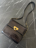 A black crocodile-embossed leather handbag with a gold-tone turn-lock clasp and a black shoulder strap, displayed on a dark gray marble surface.