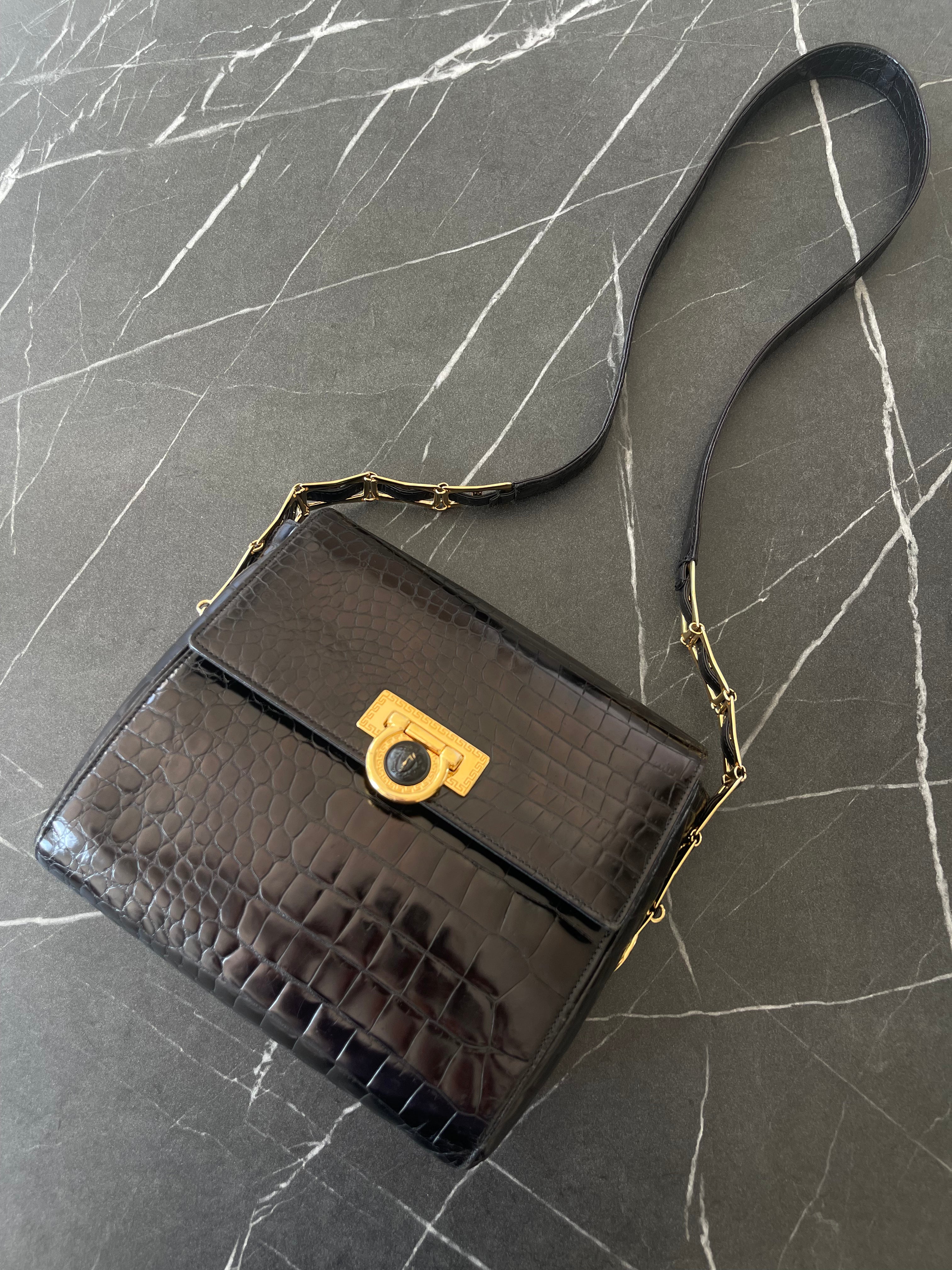 A black crocodile-embossed leather handbag with a gold-tone turn-lock clasp and a black shoulder strap, displayed on a dark gray marble surface.