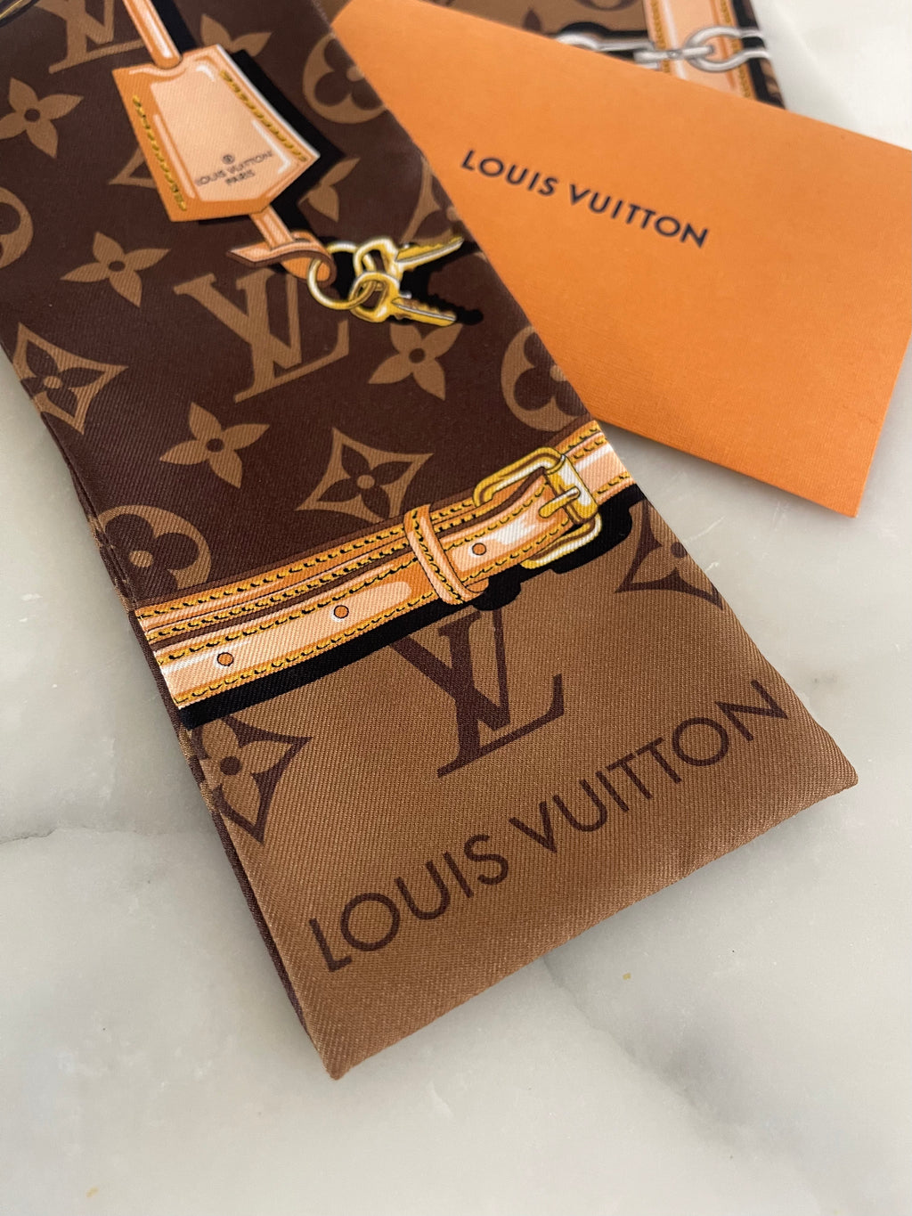 Louis Vuitton monogram scarf with a printed leather-buckle motif, shown with an orange Louis Vuitton dust bag and brand tag on a light marble surface.