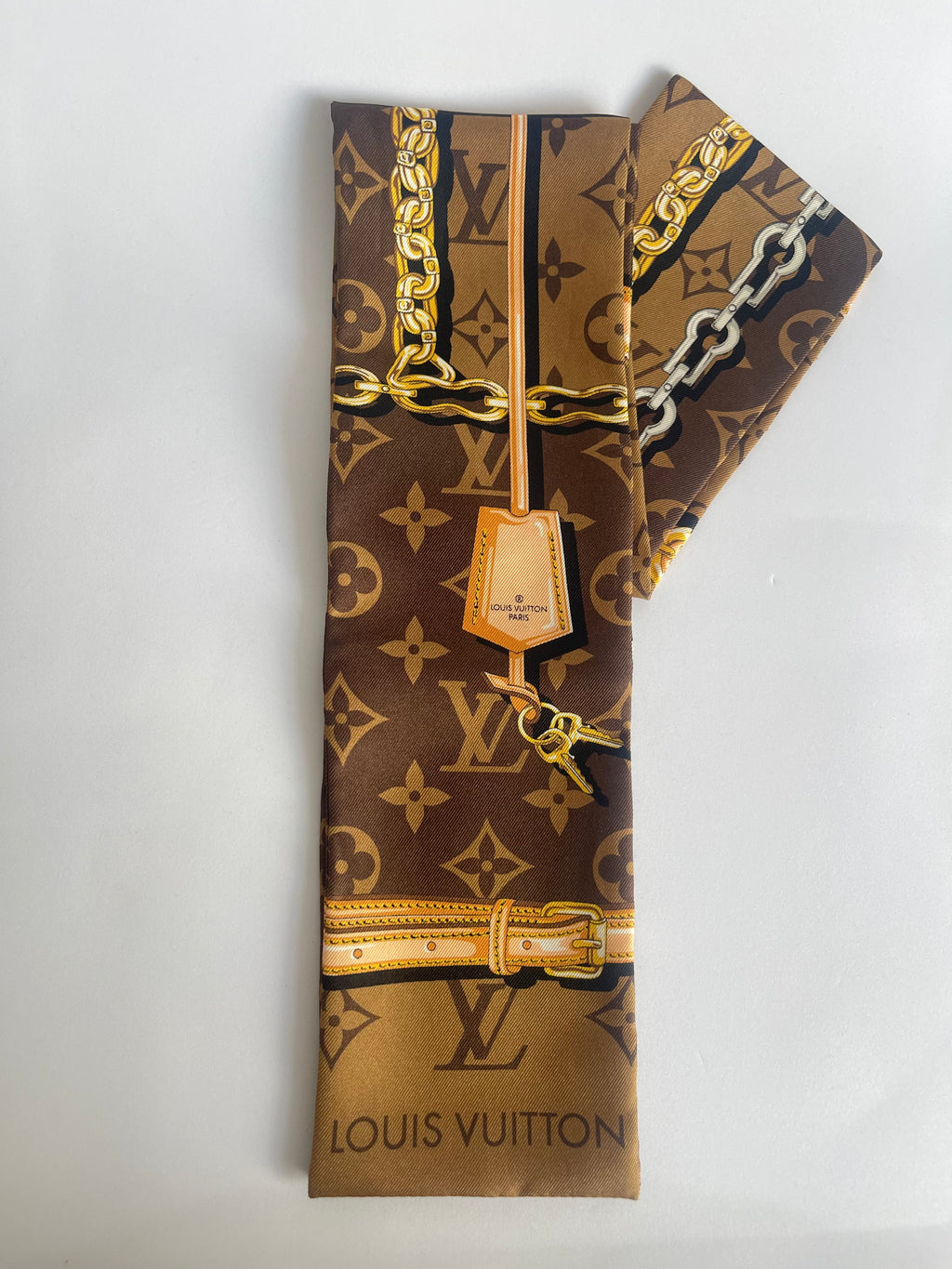 Louis Vuitton monogram silk scarf featuring a brown and gold monogram pattern with gold chain and belt graphic details, shown folded on a white background.