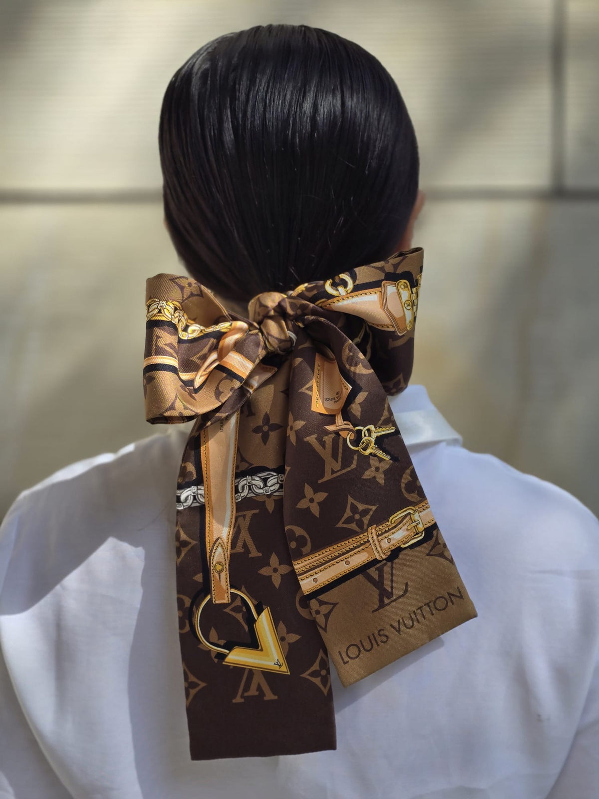 A Louis Vuitton silk scarf with a brown and gold monogram pattern, styled in a large bow around a person's dark hair, shown from behind.