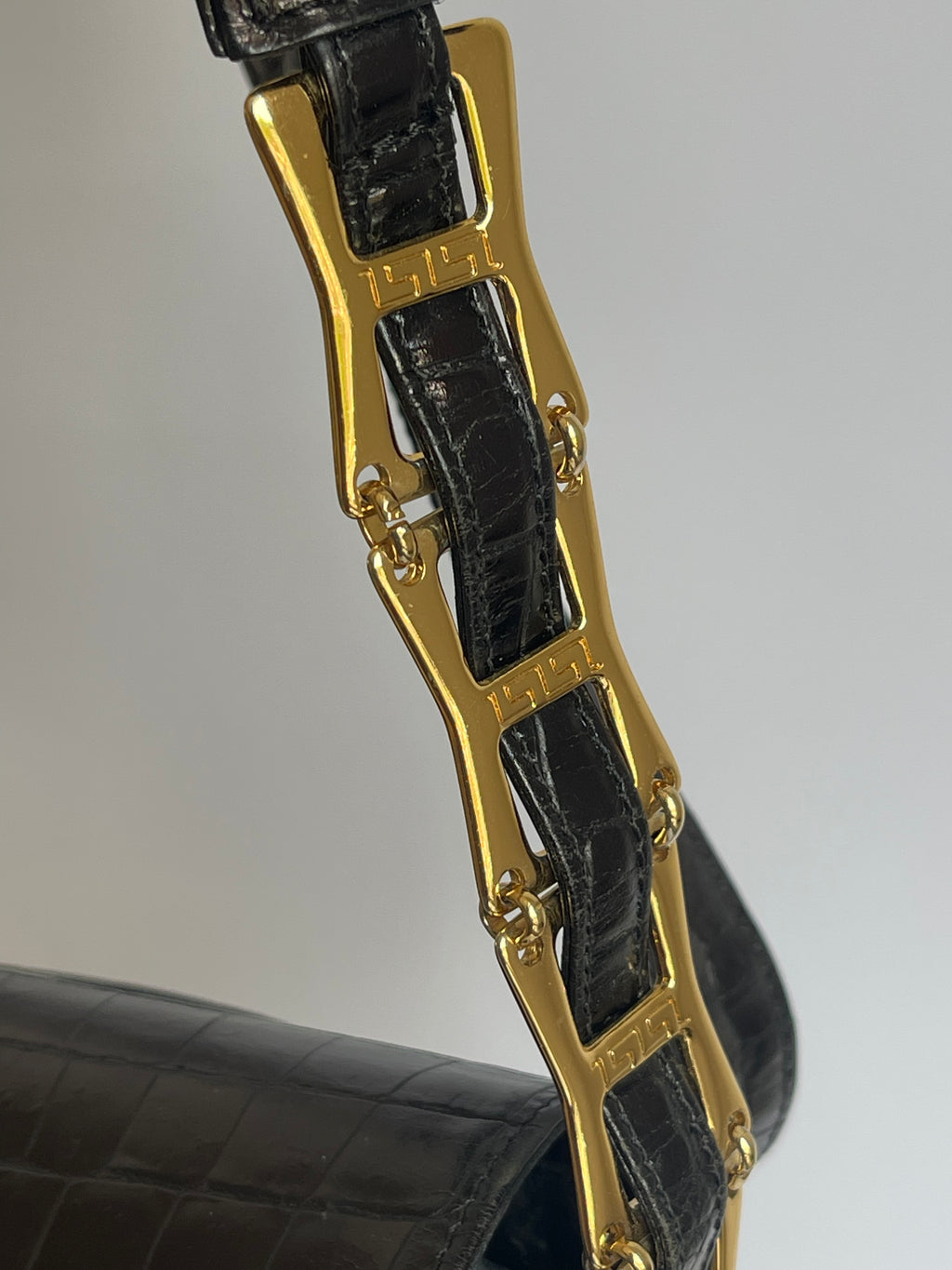 Close-up of a black crocodile-embossed leather strap with a prominent gold-tone metal link clasp engraved with the brand name 'Versace'.
