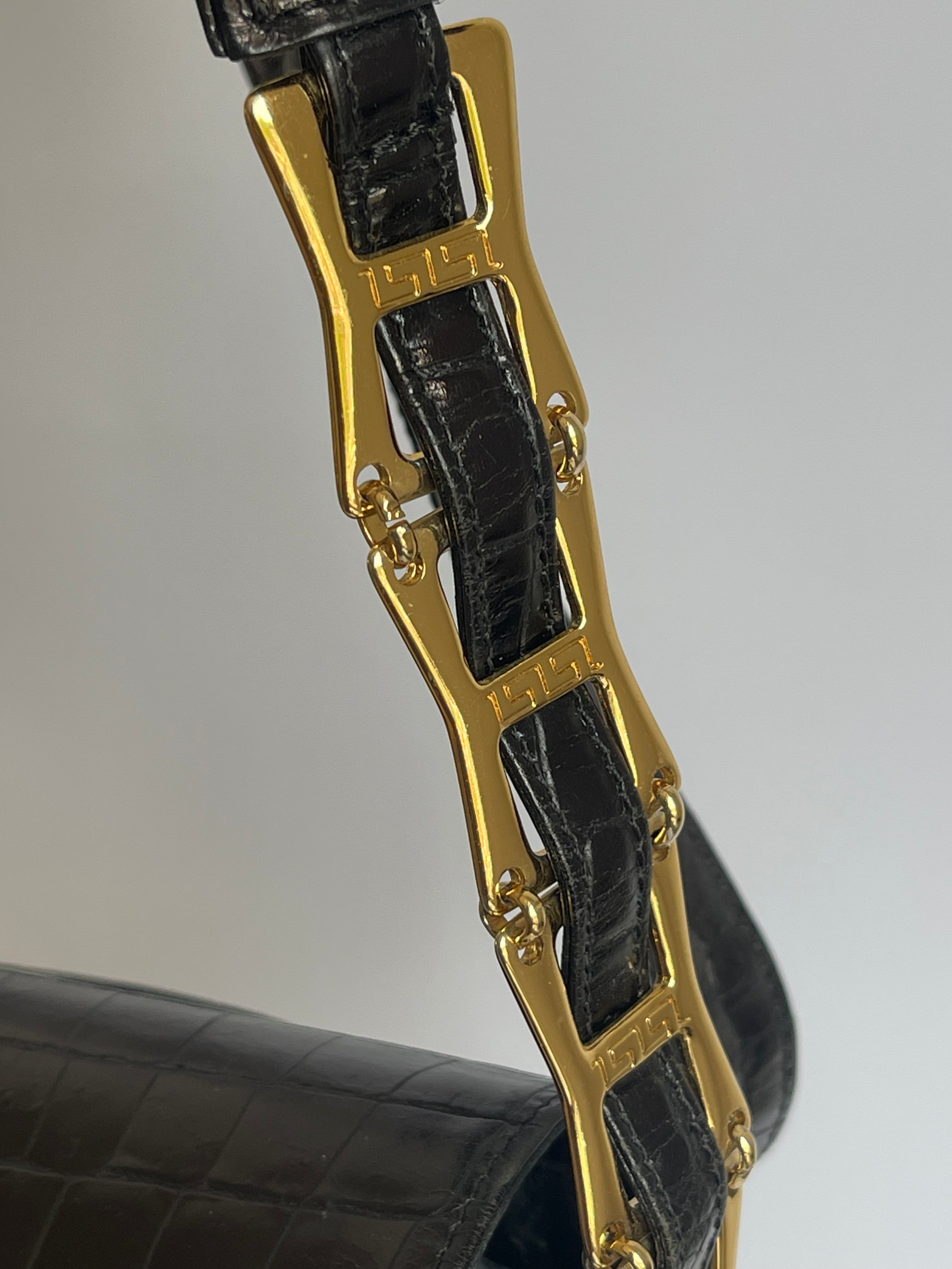 Close-up of a black crocodile-embossed leather strap with a prominent gold-tone metal link clasp engraved with the brand name 'Versace'.