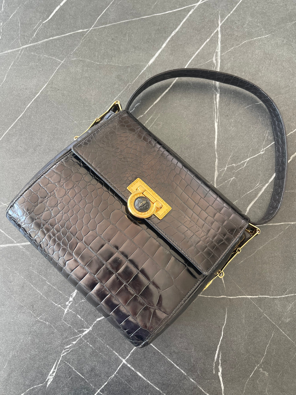 A structured black crocodile-embossed leather handbag with a top handle and a gold-tone turn-lock clasp, displayed on a gray marble-patterned surface.