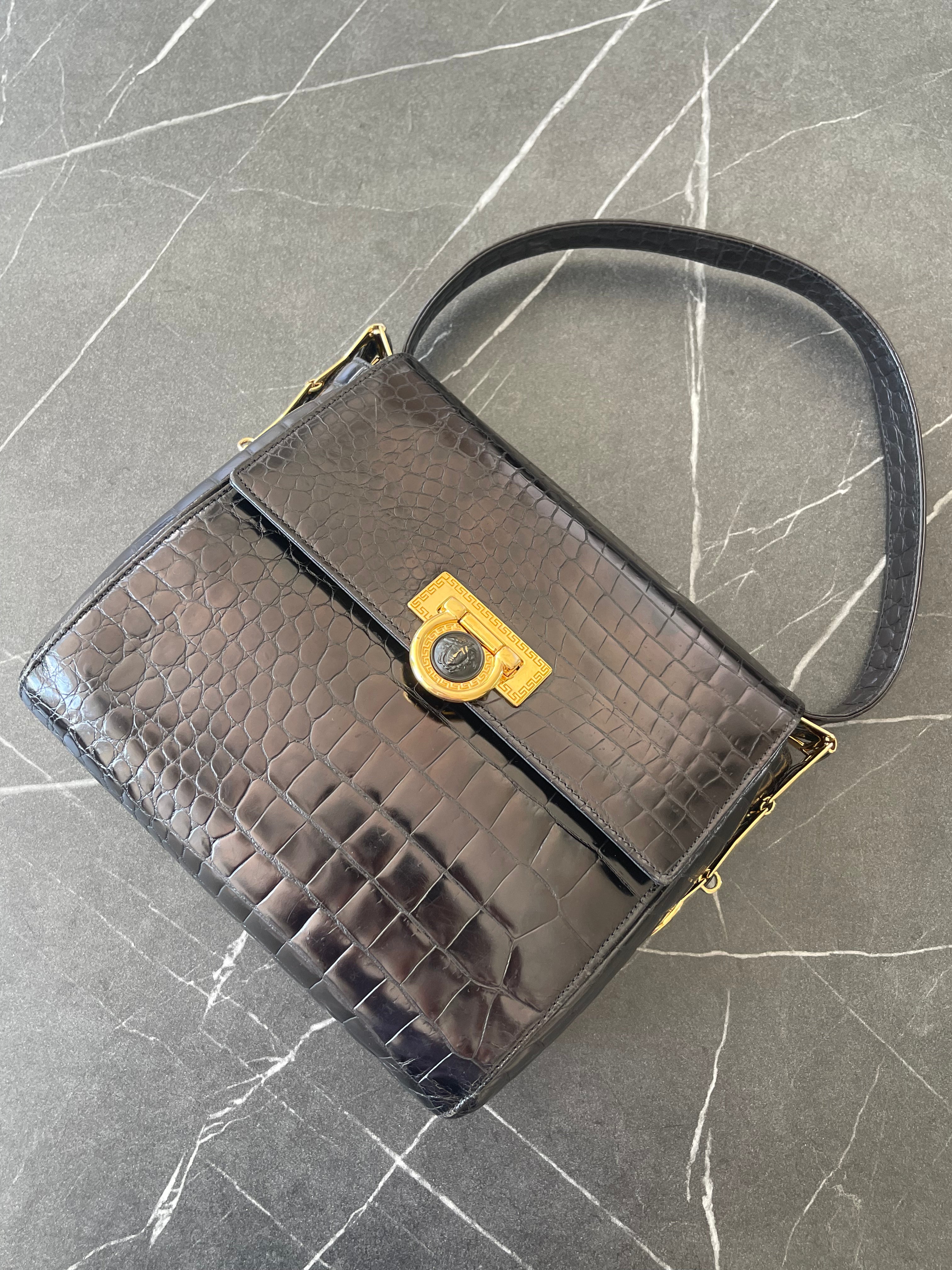 A structured black crocodile-embossed leather handbag with a top handle and a gold-tone turn-lock clasp, displayed on a gray marble-patterned surface.
