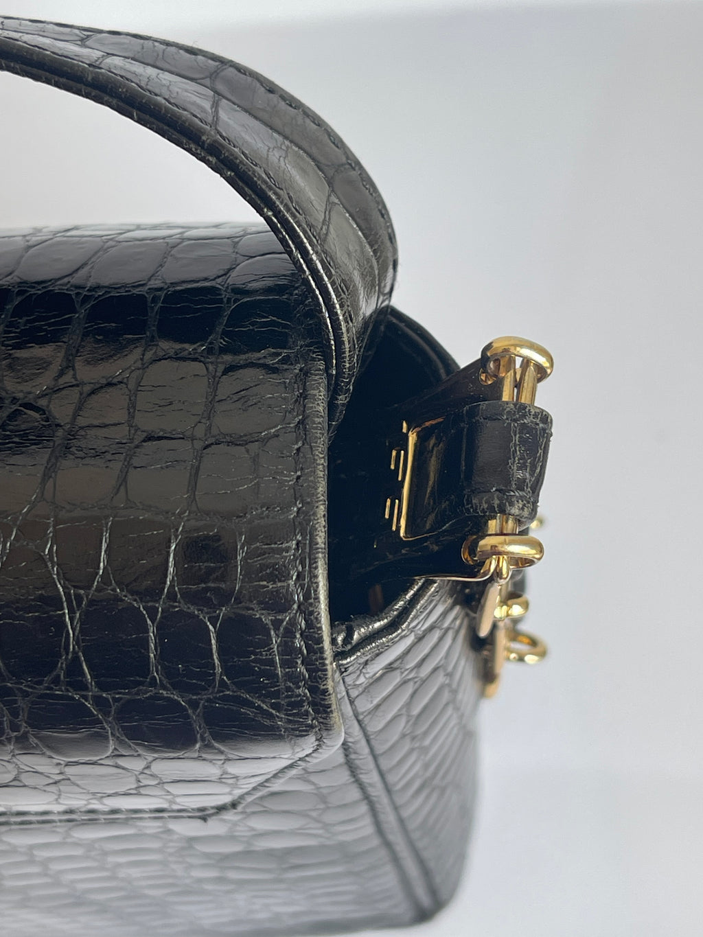 Close-up of a black crocodile-embossed leather handbag showing a gold-tone metal clasp and part of the strap on a light gray background.