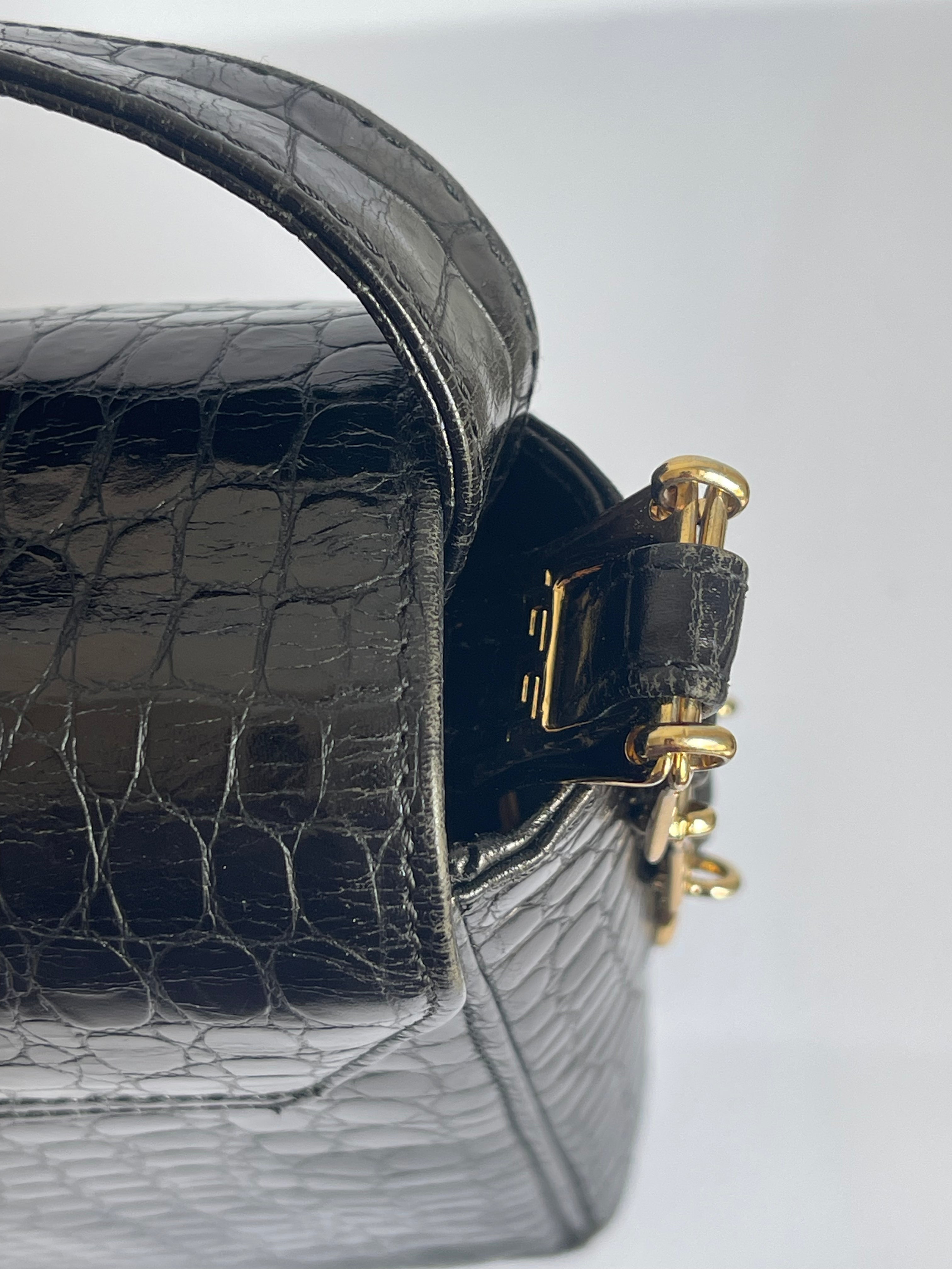 Close-up of a black crocodile-embossed leather handbag showing a gold-tone metal clasp and part of the strap on a light gray background.