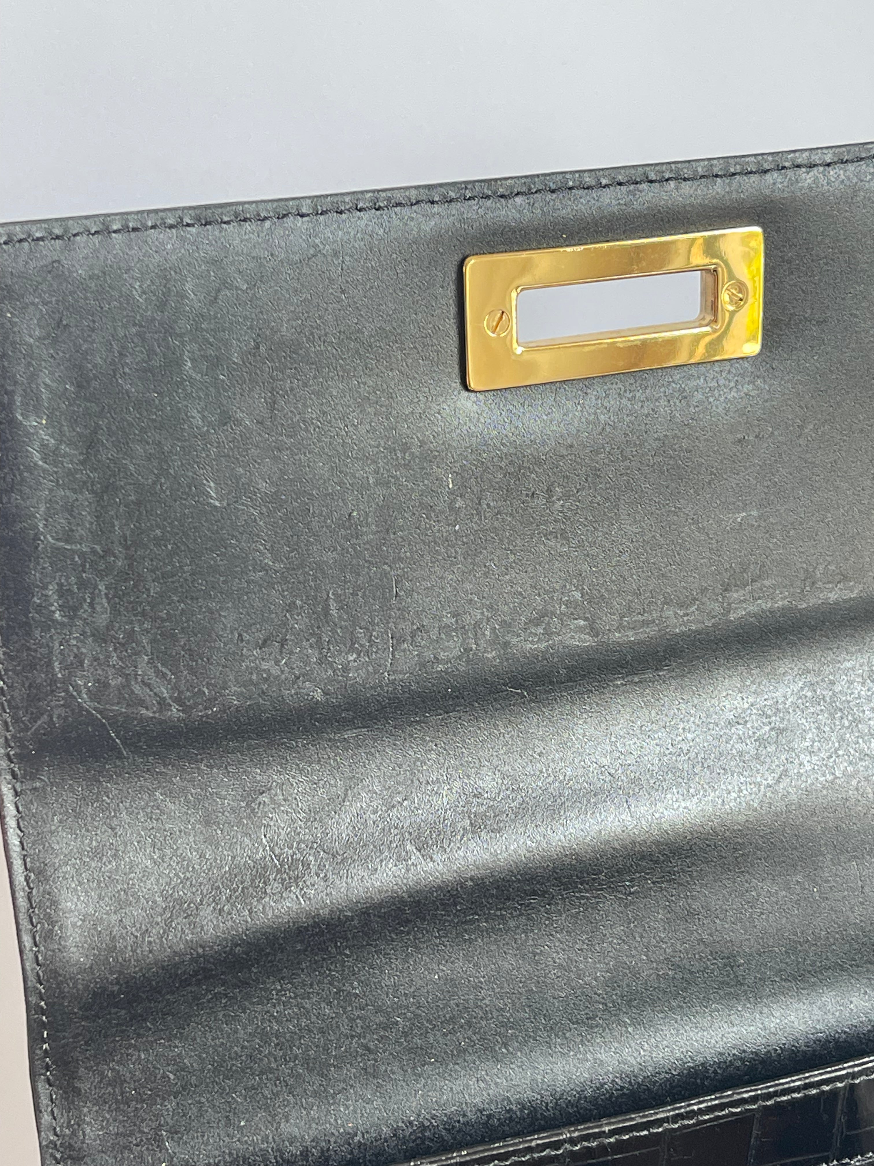 Close-up of a black leather clutch or wallet featuring a prominent gold-tone metal slot clasp on the front flap.