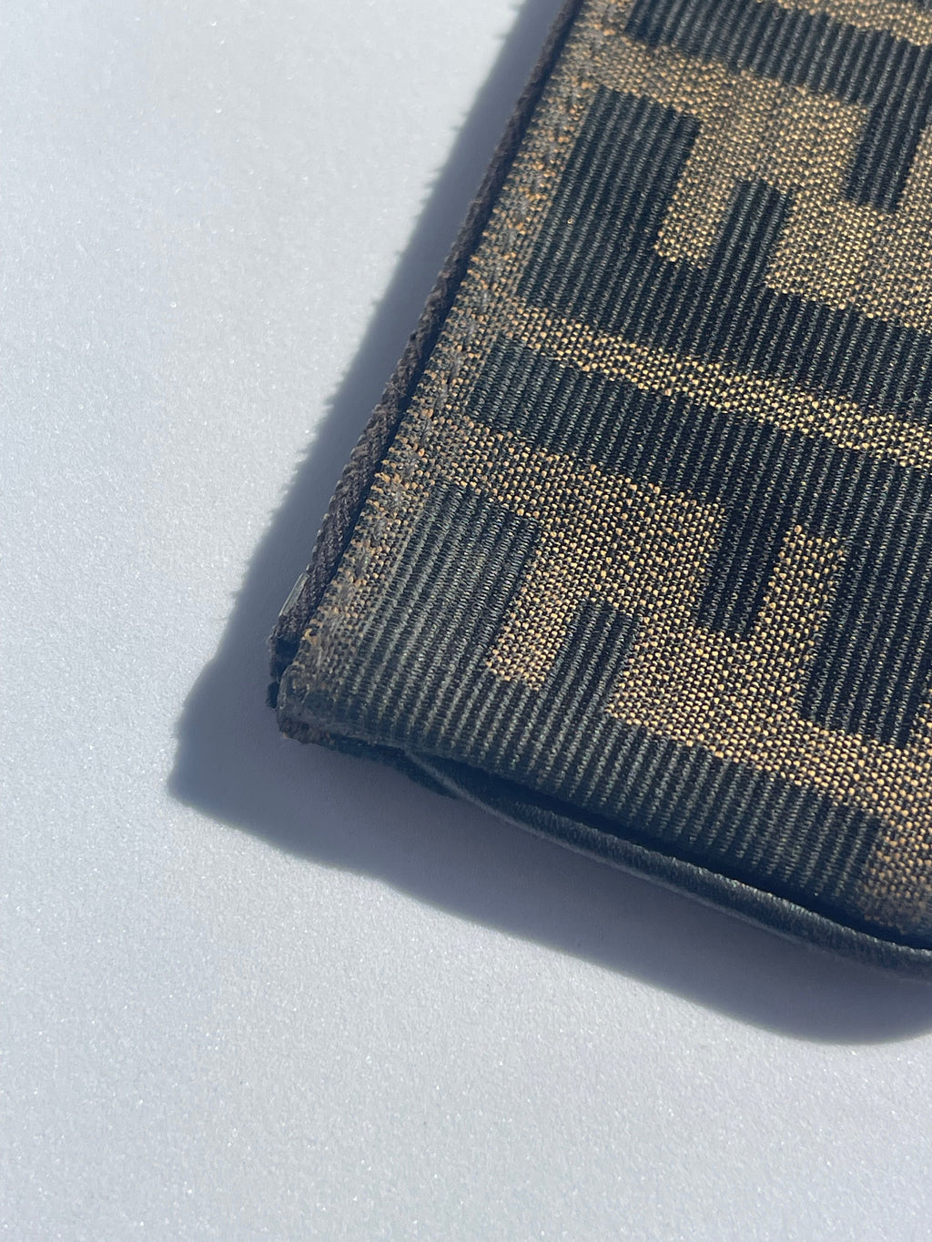 A close-up of a Fendi wallet in the signature Zucca monogram canvas, featuring a dark brown and black woven pattern, resting on a light gray surface with a sharp shadow.