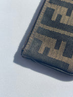 A close-up of a Fendi wallet in the signature Zucca monogram canvas, featuring a dark brown and black woven pattern, resting on a light gray surface with a sharp shadow.