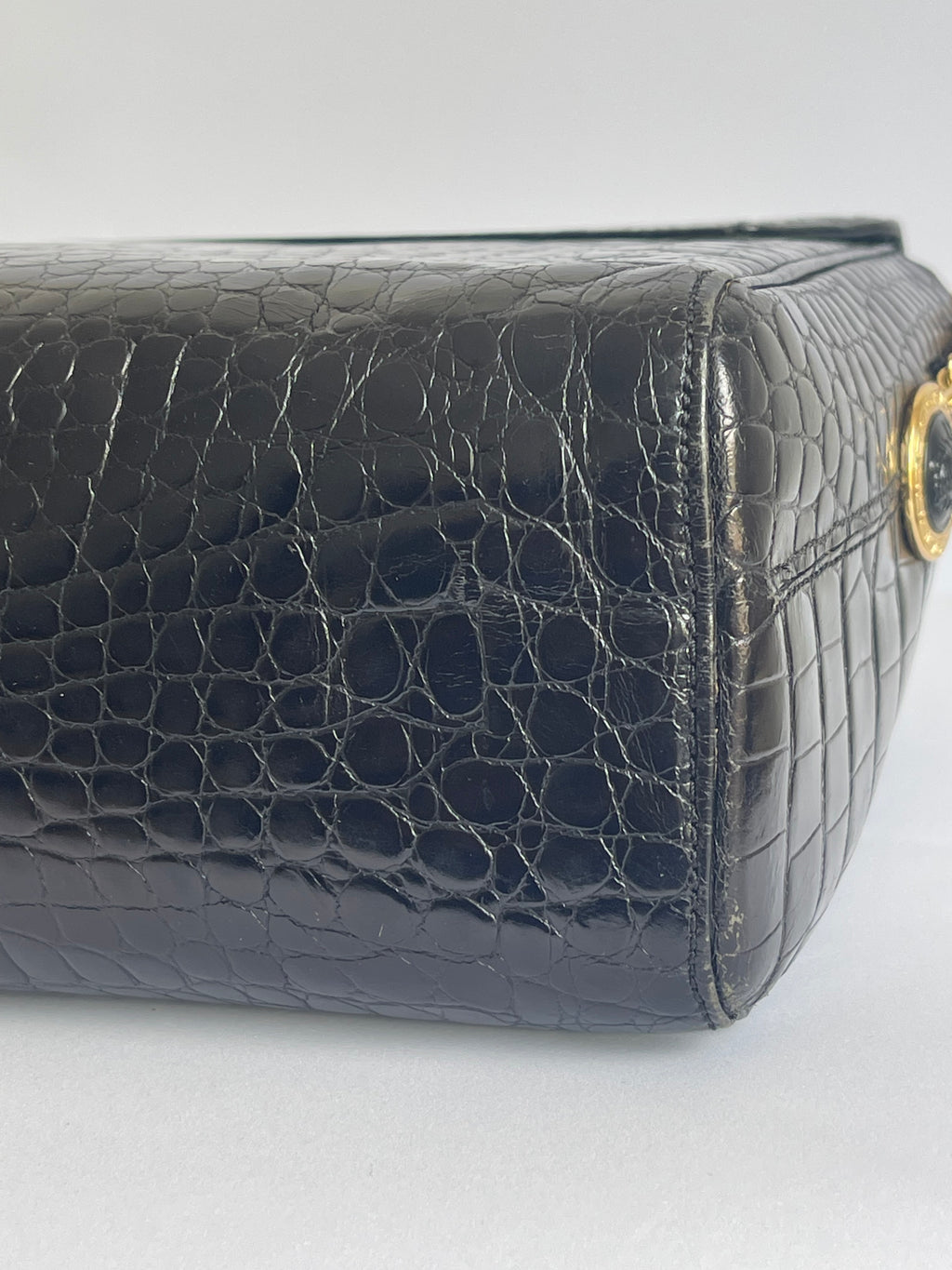 Close-up of a black crocodile-embossed leather handbag corner showing a gold-tone ring detail on a light gray background.