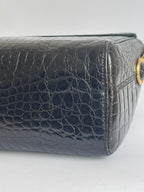 Close-up of a black crocodile-embossed leather handbag corner showing a gold-tone ring detail on a light gray background.