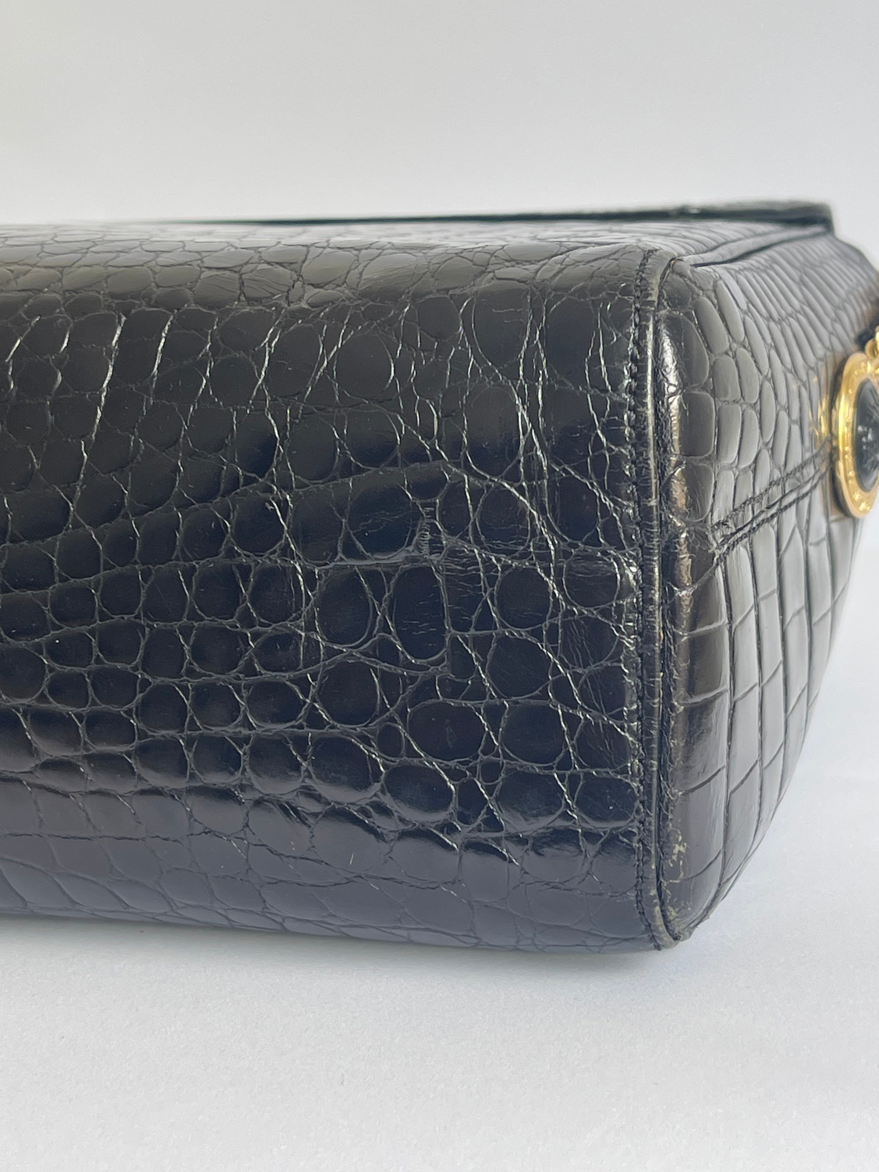 Close-up of a black crocodile-embossed leather handbag corner showing a gold-tone ring detail on a light gray background.