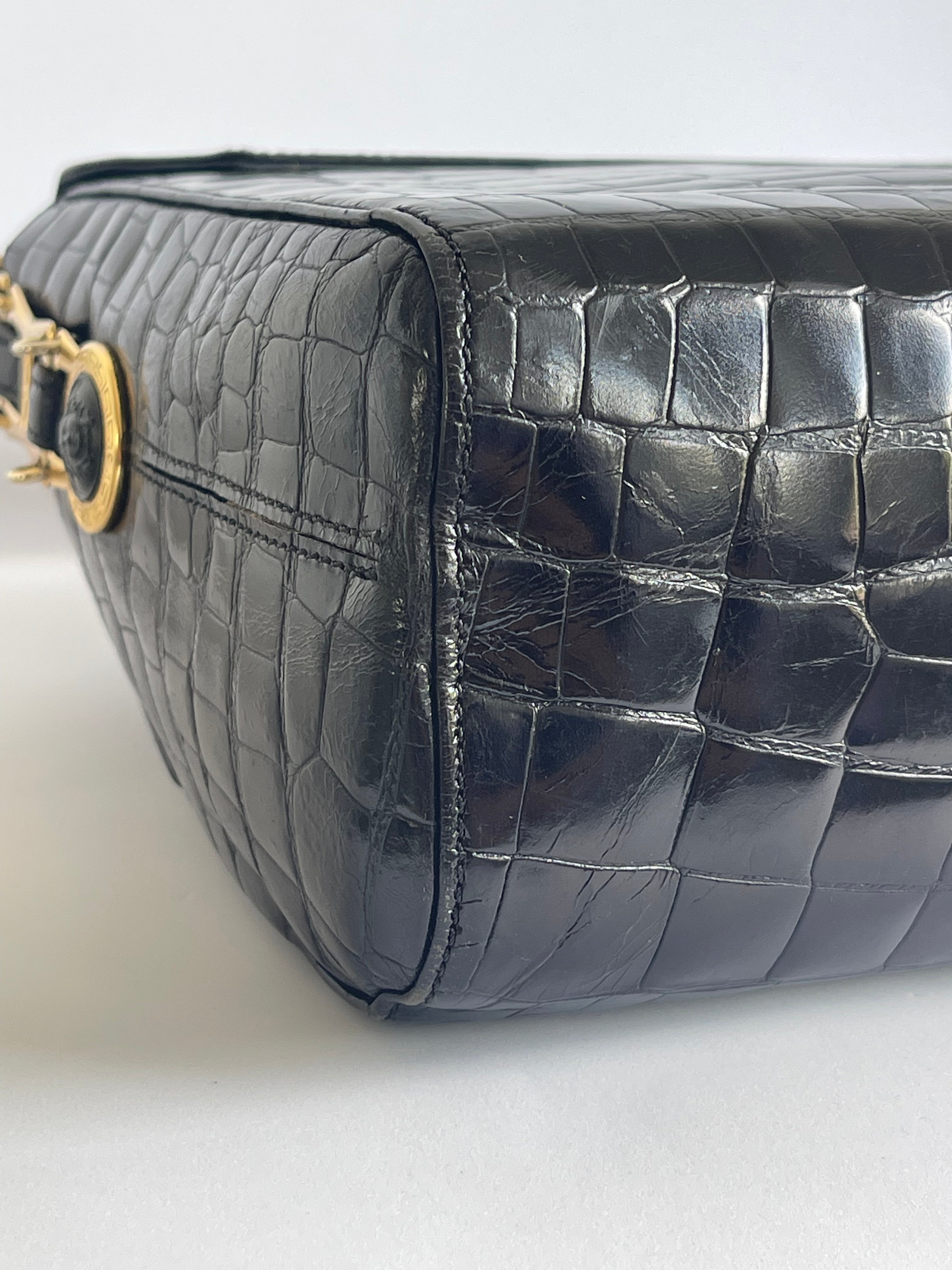 Close-up of a black crocodile-embossed leather handbag showing the side profile and a gold-tone hardware detail on a light gray background.