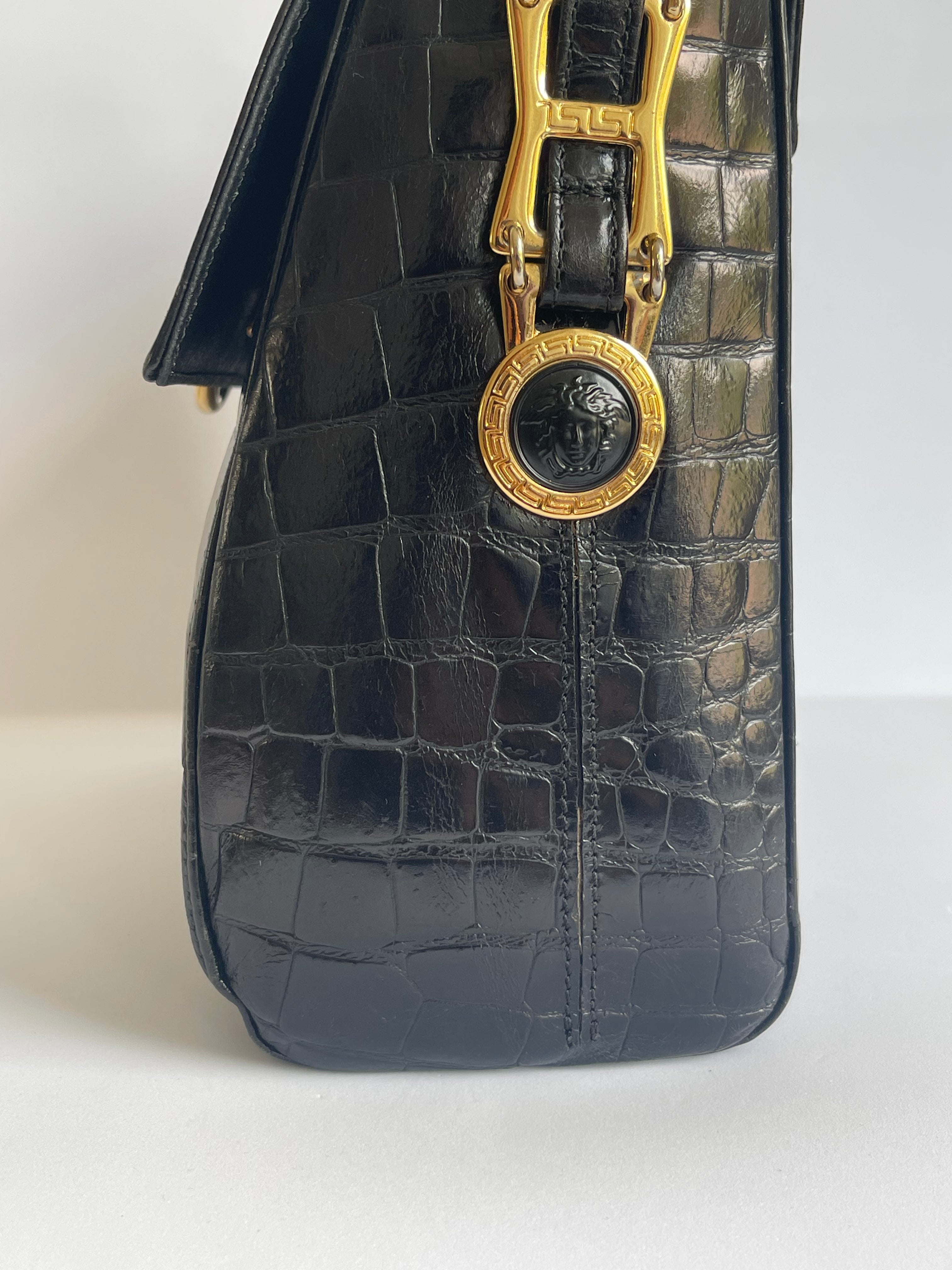 Close-up of a black crocodile-embossed leather handbag featuring gold-tone hardware and a circular medallion clasp with a monogram design, set against a plain white background.